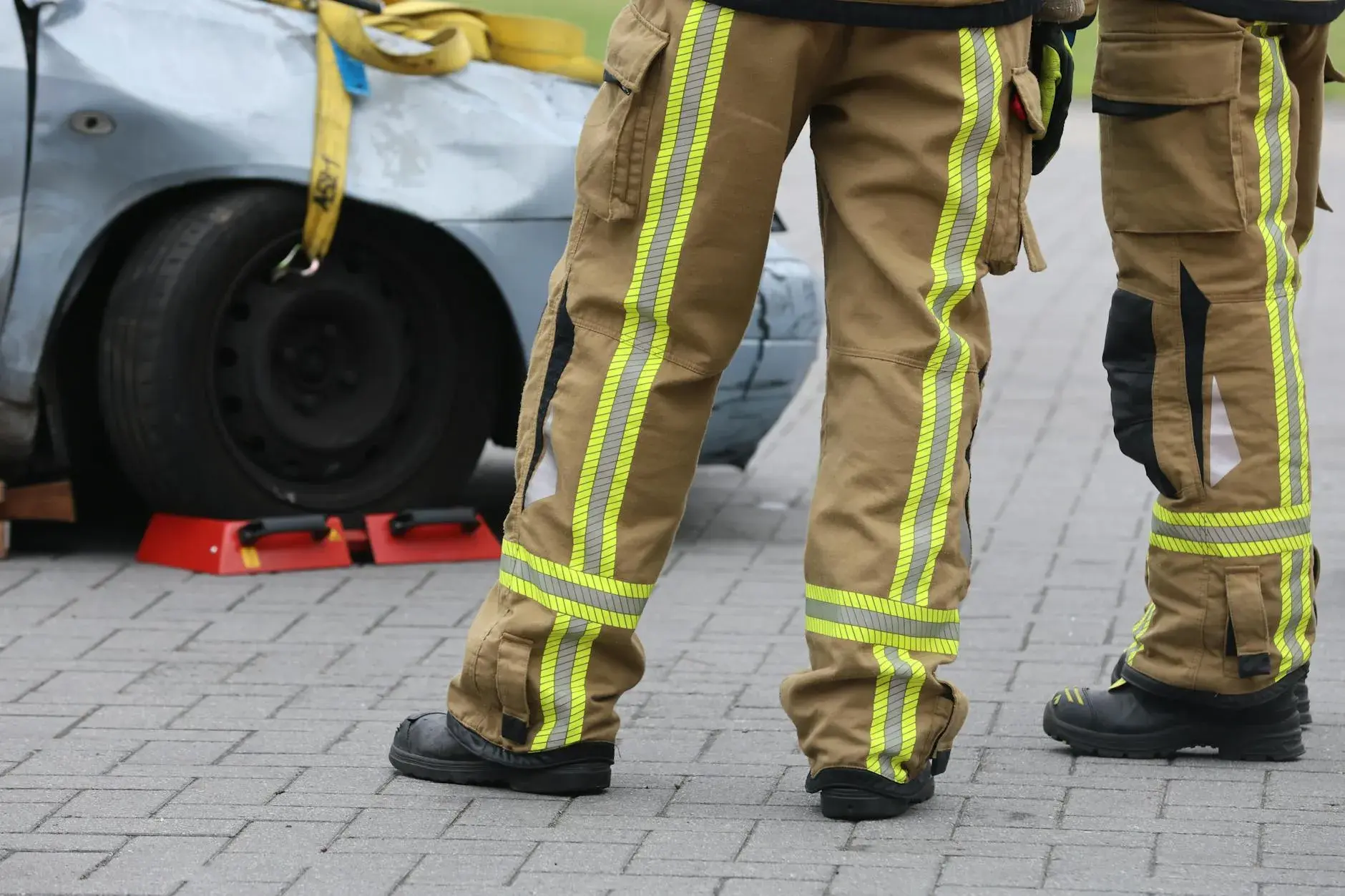 Brandmænd inspicerer en bilulykke iført beskyttelsesudstyr. Fokus er på deres bukser og fodtøj under arbejdet på stedet.
