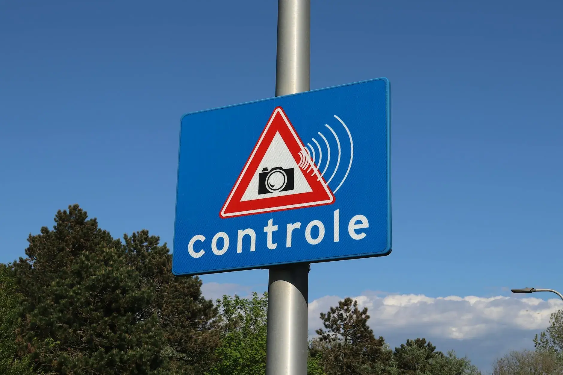 Et blåt vejskilt med et kamera-ikon og teksten “controle” markerer, at der foretages hastighedskontrol. Skiltet signalerer overvågning langs vejen.