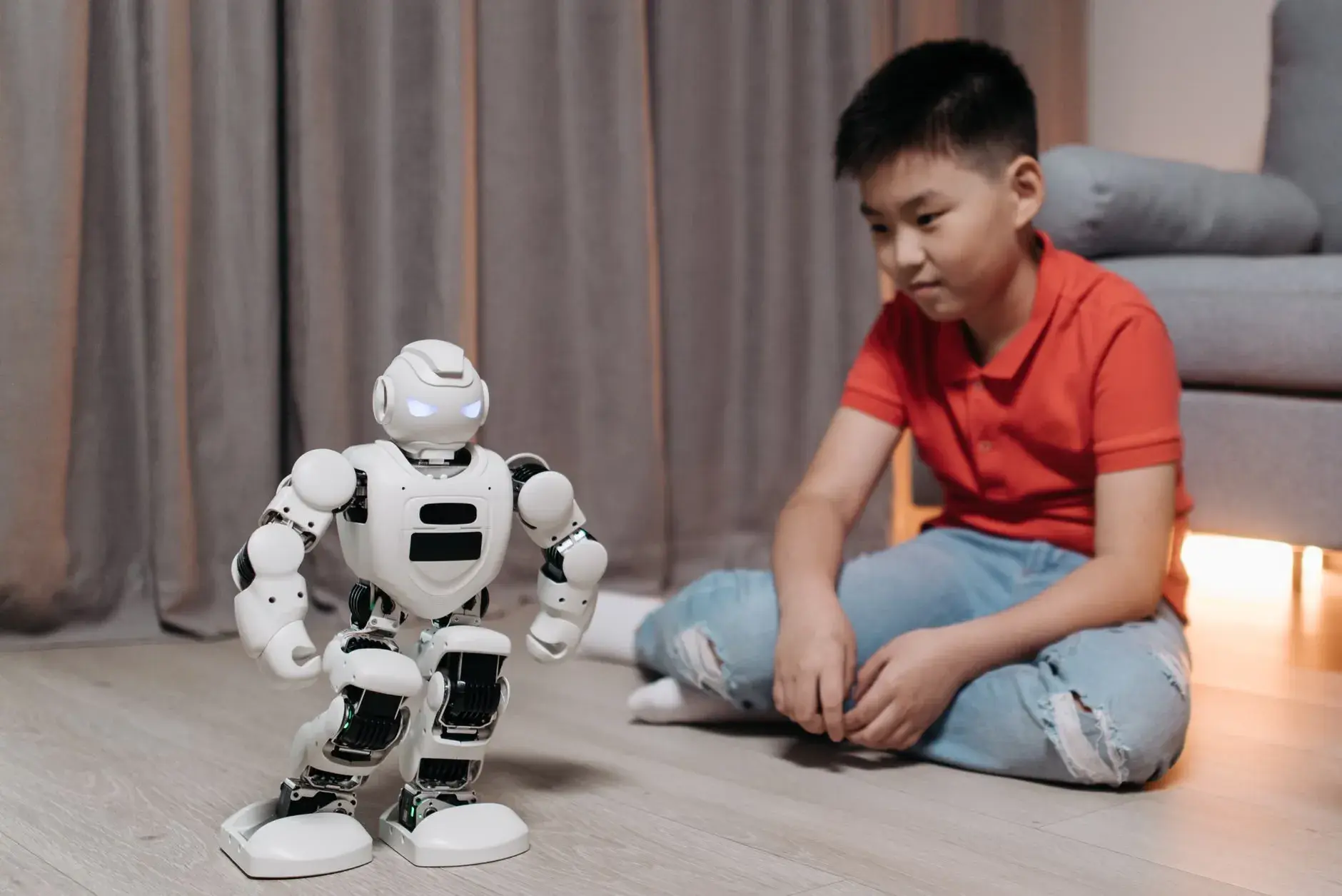 En asiatisk dreng sidder på gulvet og interagerer med en hvid robot, der viser innovation og teknologi.