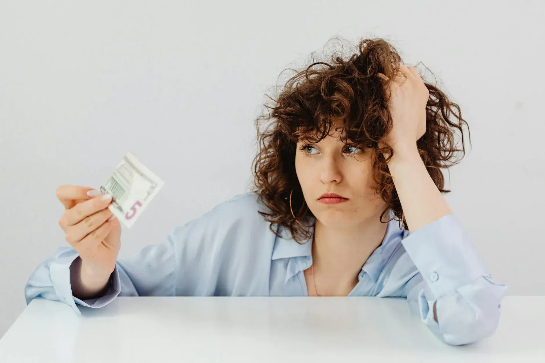 En bekymret kvinde med krøllet hår holder en femdollarseddel og udtrykker økonomisk uro. Billedet formidler stress og bekymring omkring penge.