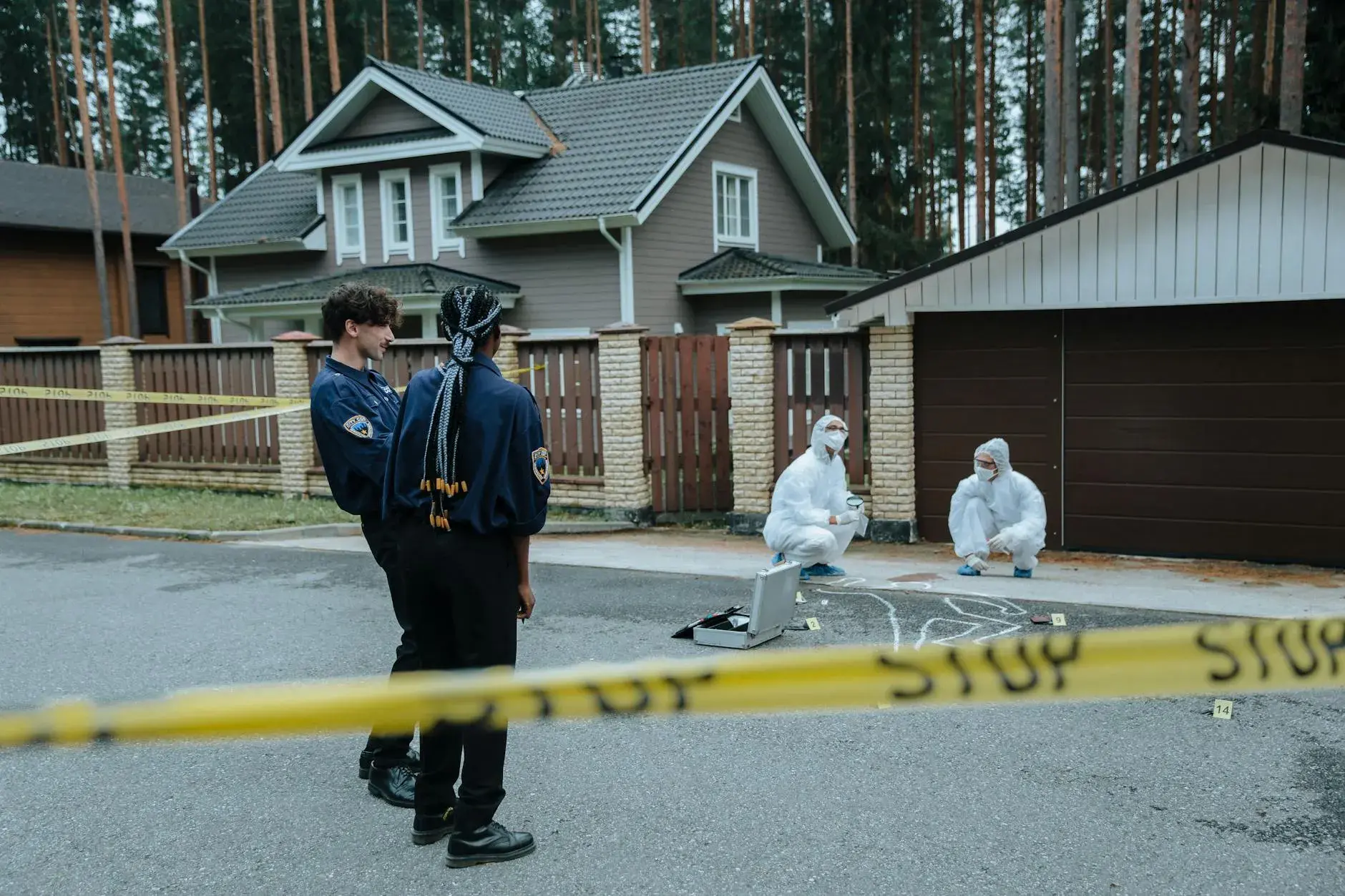 Politi og forensic-eksperter undersøger et gerningssted foran parcelhuse. Scenen er dokumenteret som led i en efterforskning.