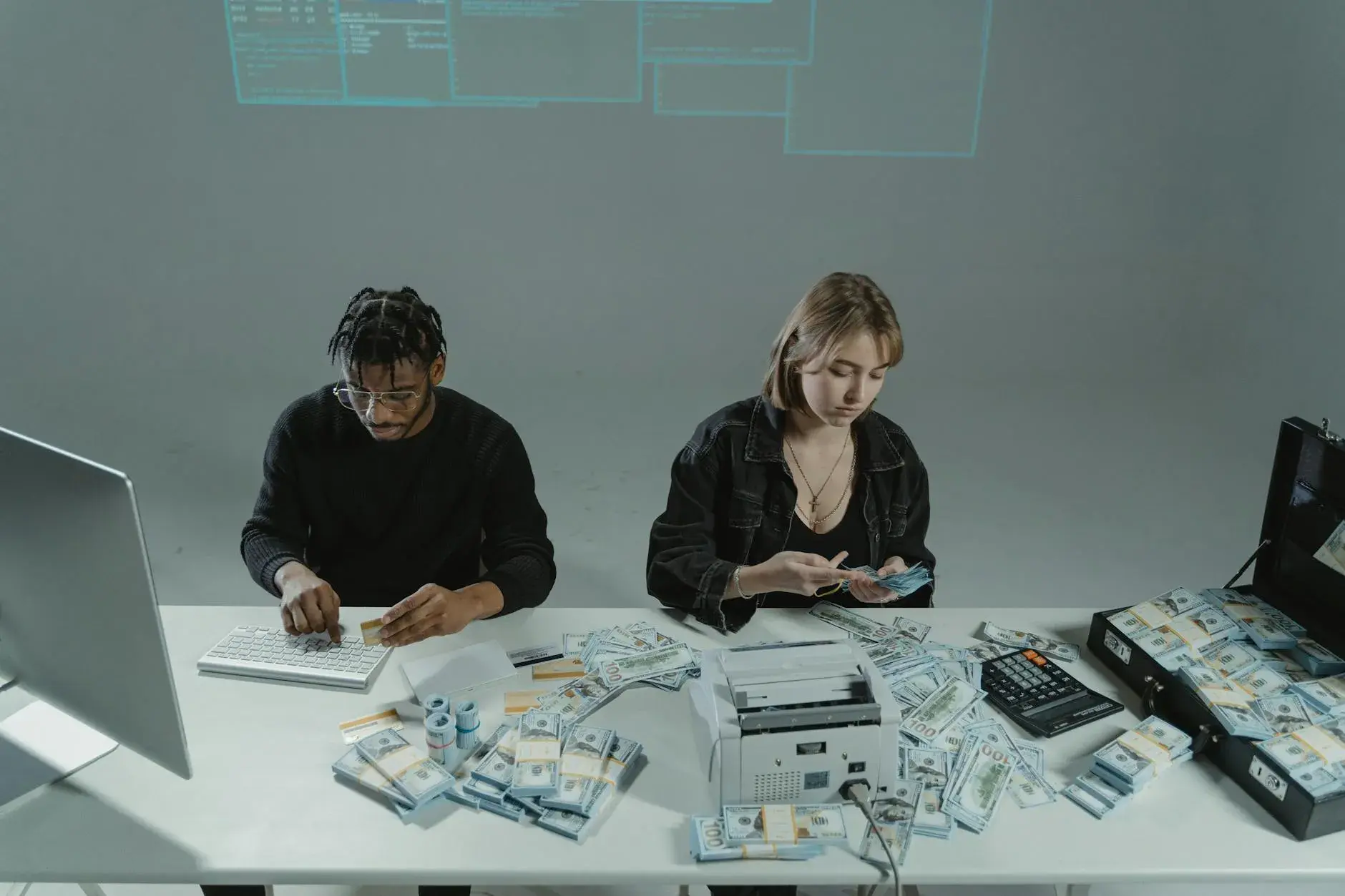 To personer tæller penge ved et skrivebord med computere og kontantudstyr. Billedet formidler arbejde med økonomi, betalinger eller kassehåndtering.