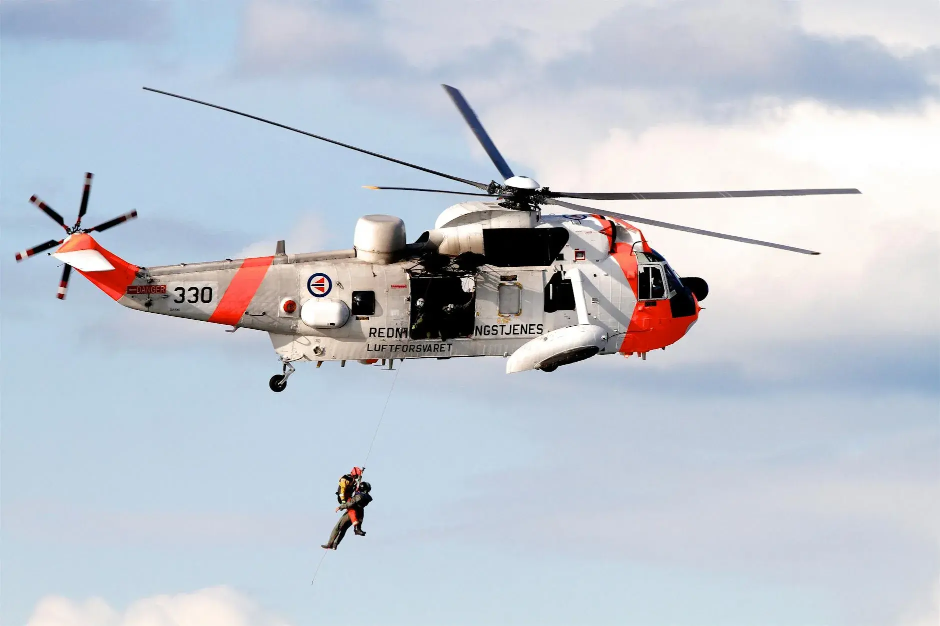 En redningshelikopter fra Royal Norwegian Air Force udfører en mid-air hoist-operation på en klar dag. Scenen viser redningsberedskab i luften.