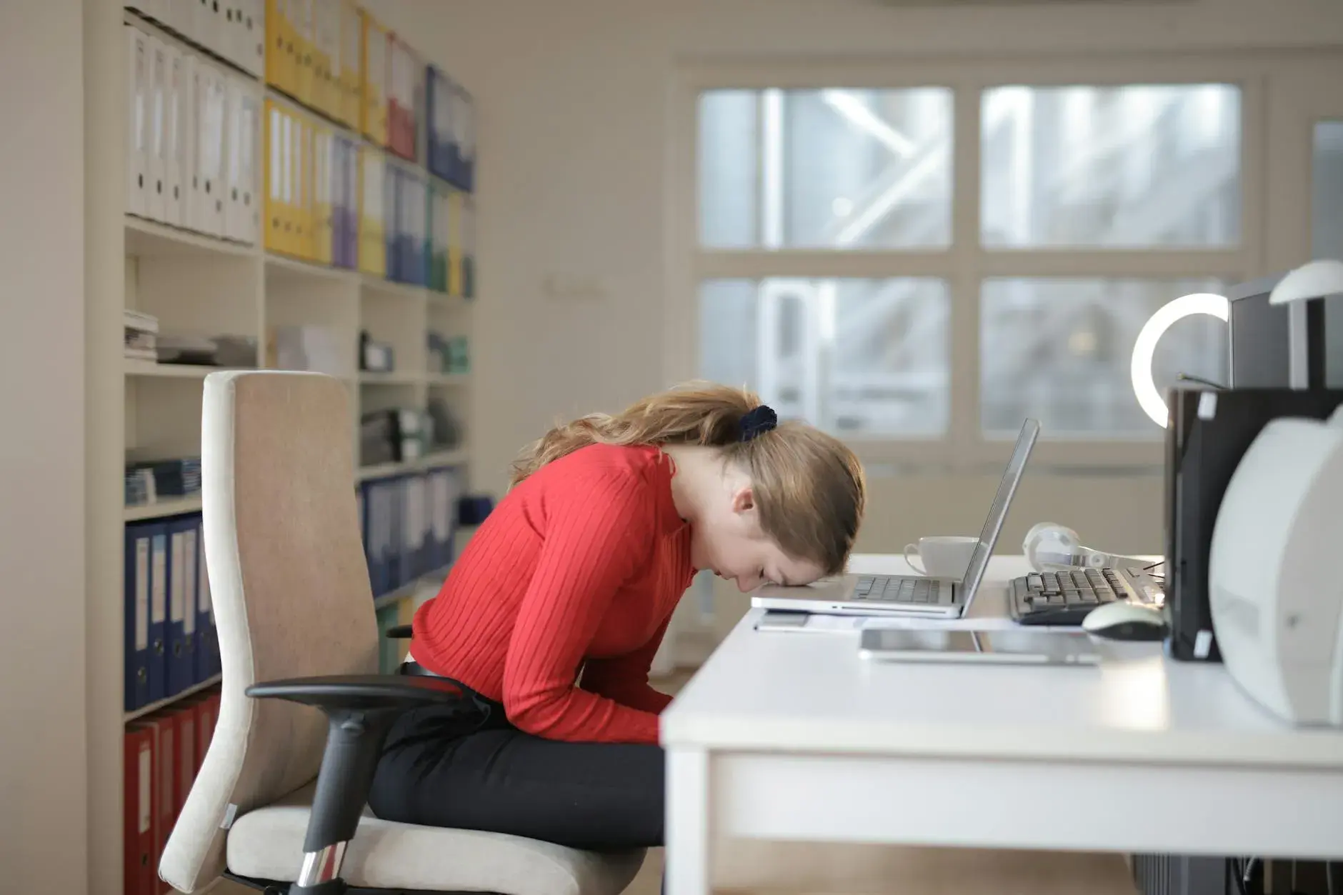 En træt kvinde i en rød sweater falder i søvn på et kontorskrivebord ved siden af en laptop. Billedet formidler overvældelse og udmattelse fra fjernarbejde.