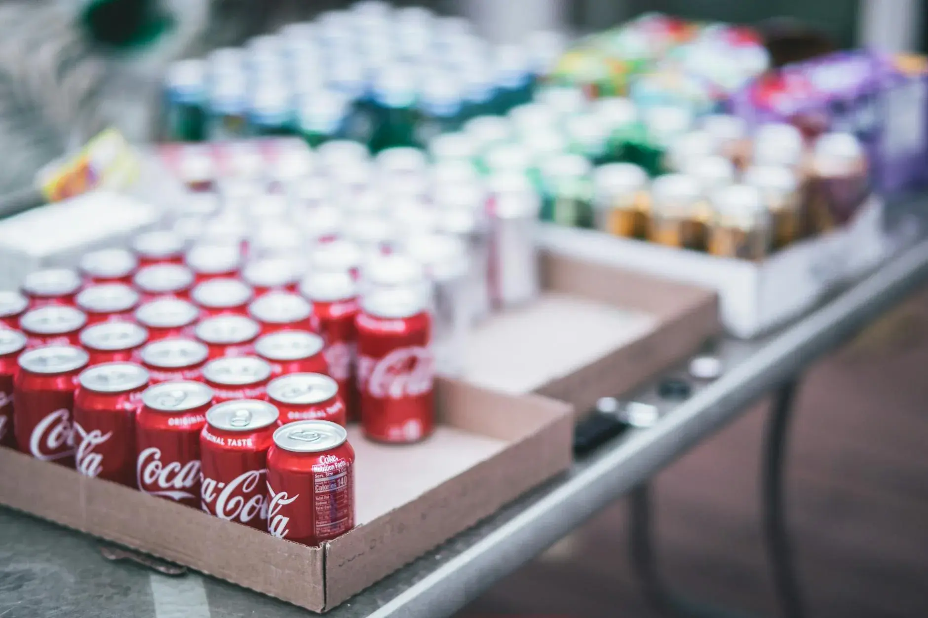 Sløret foto af en række forskellige sodavandsdåser på et bord, med Coca-Cola i fokus. Stemningen er reklameklar og passer til artikler om drikkevarer og branding.