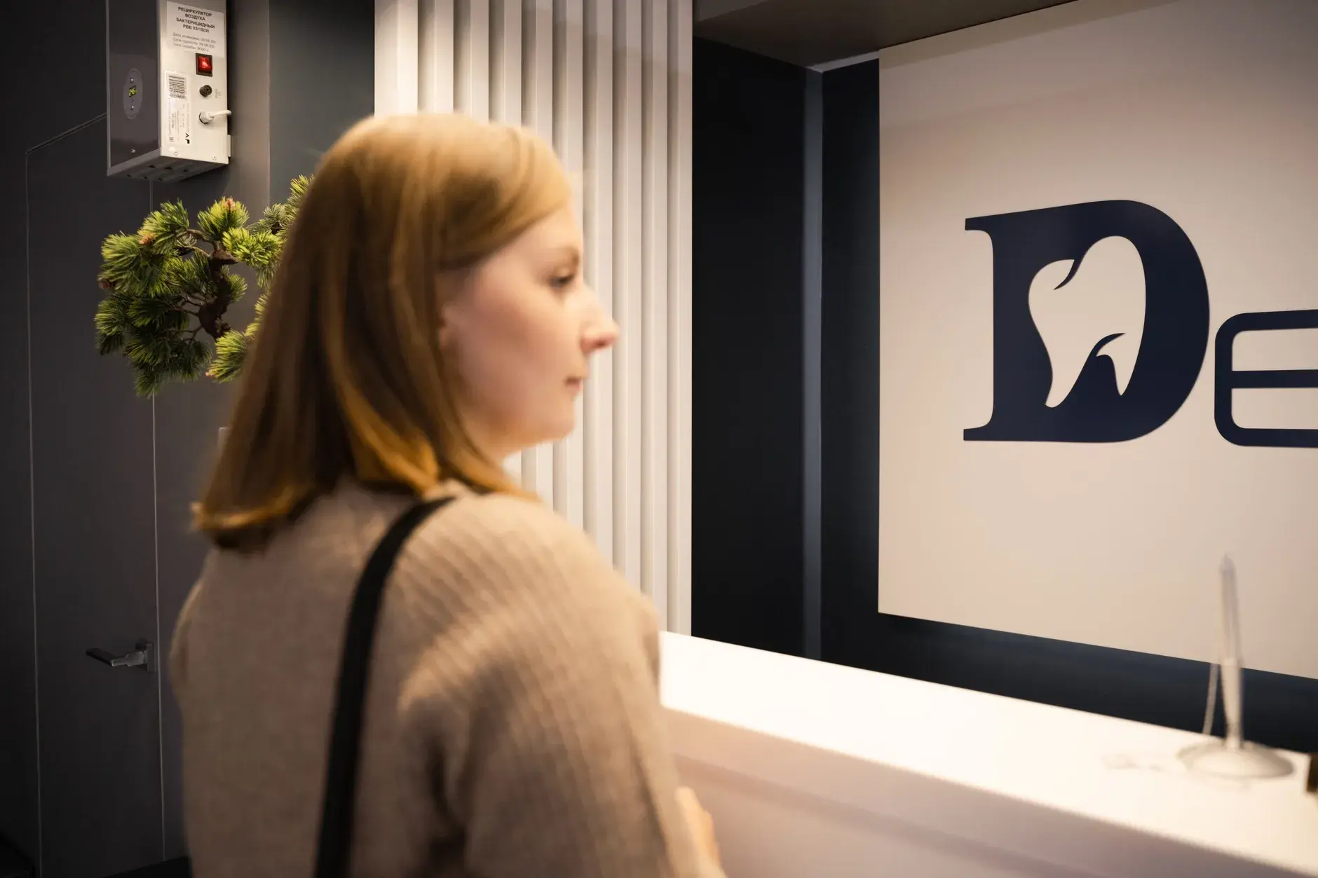En ung kvinde står ved receptionen i en tandlægeklinik i sideprofil. Stemningen er rolig og hverdagsnær.