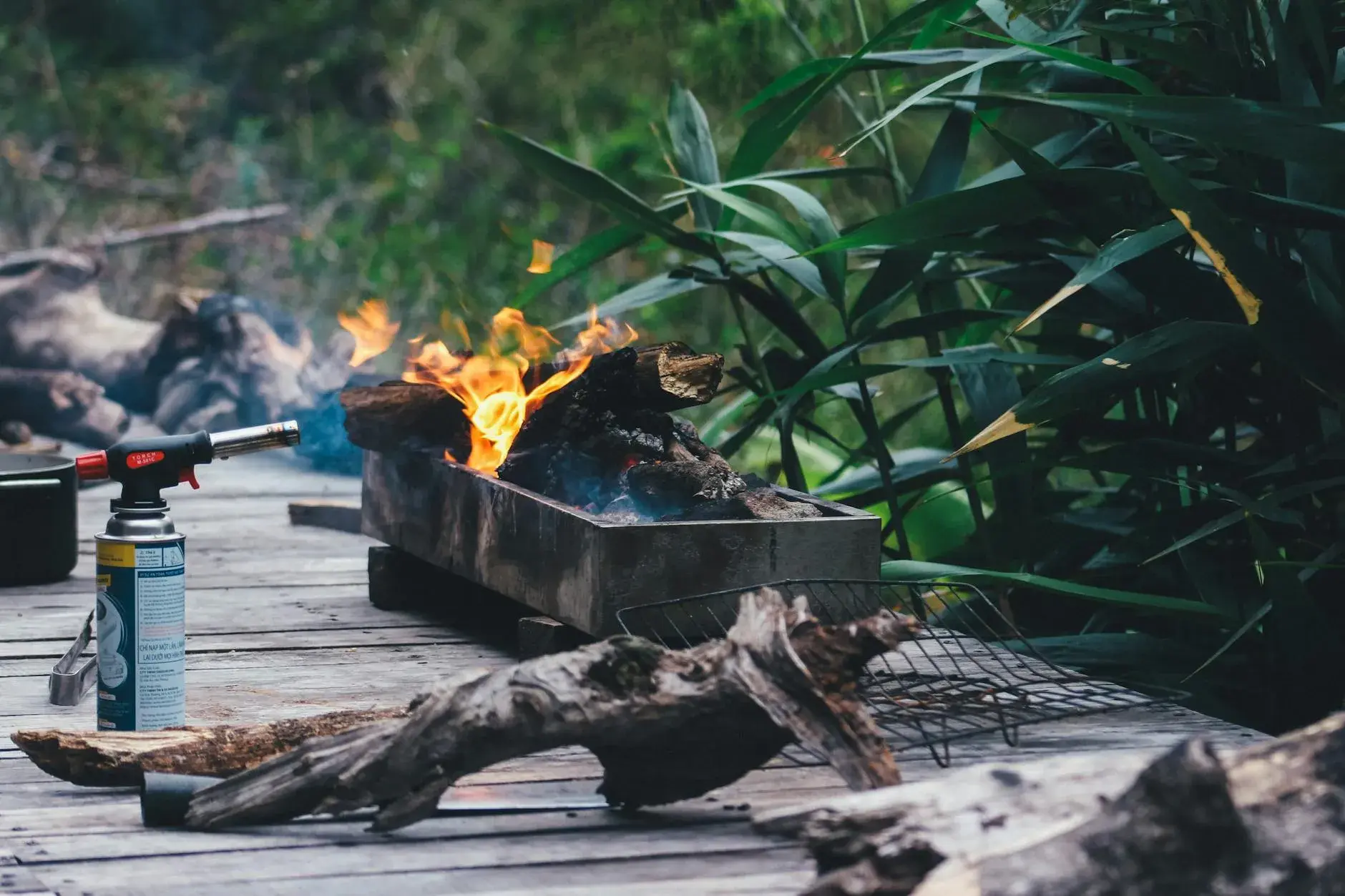 En rustik udendørs grillopsætning med brændestykker og synlige flammer i frodig, grøn beplantning. Stemningen er varm og rå, som taget fra en hyggelig haveaften.