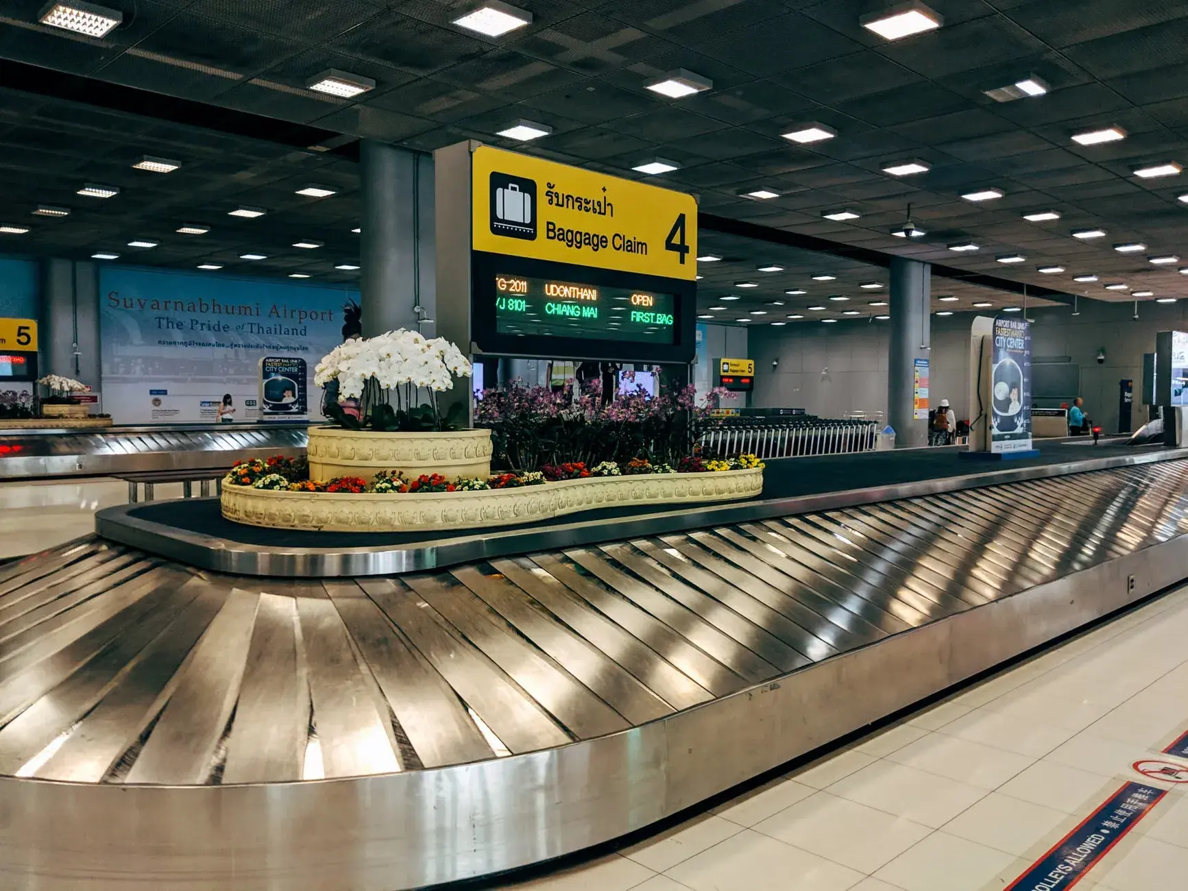 Et moderne bagageafhentningsområde på Suvarnabhumi Lufthavn i Bangkok med bagagekarusel og skilte. Rejsende samles omkring for at hente deres bagage.