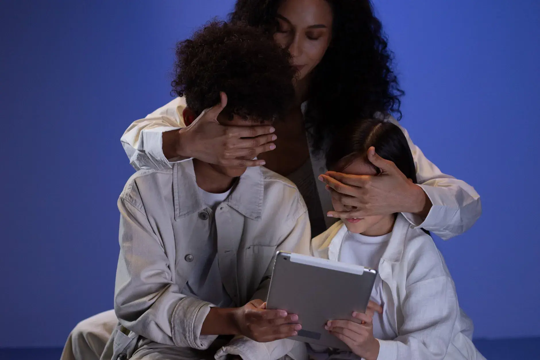 En mor dækker sine børns øjne, mens de holder en tablet, som en illustration af forældrekontrol. Billedet formidler beskyttelse og grænsesætning i digitale medier.