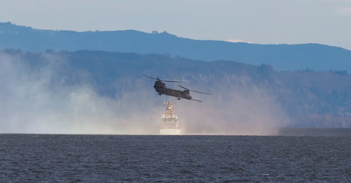 A military helicopter flying low over water near a ship with forested mountains behind it.