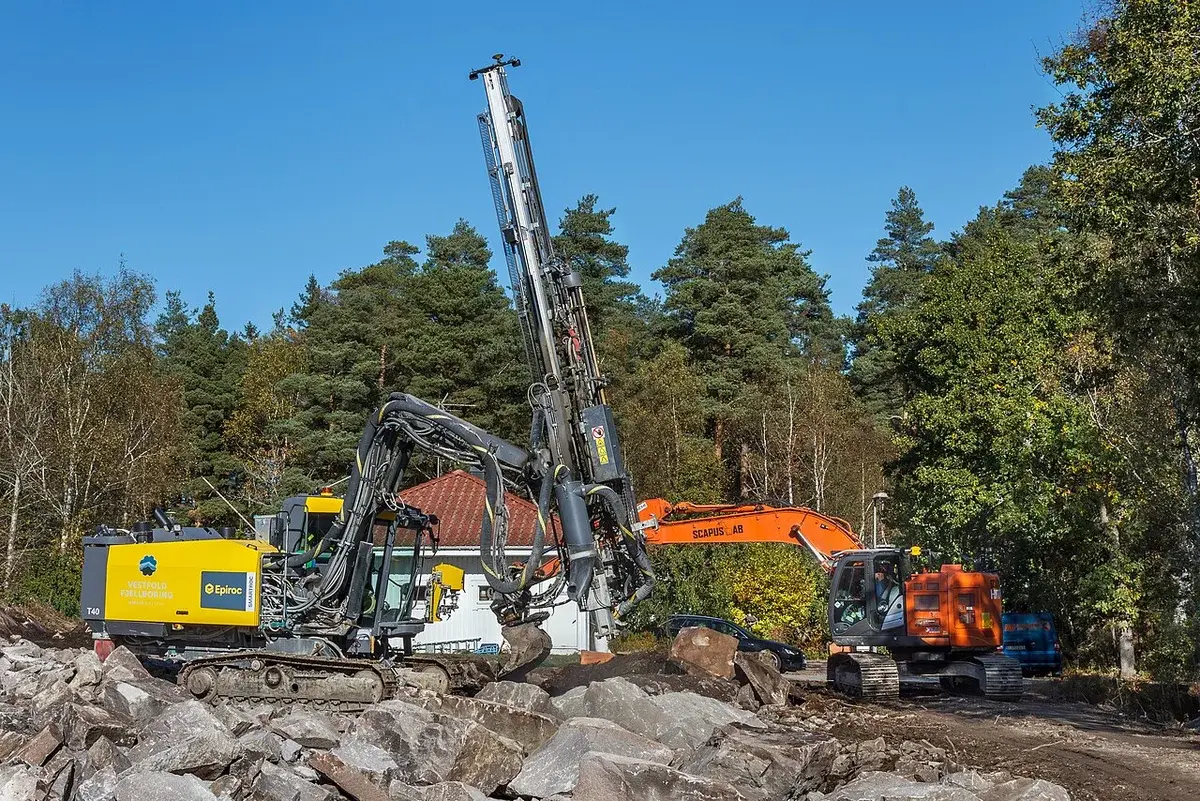 A drilling rig and an excavator working at a rocky construction site in Sweden.