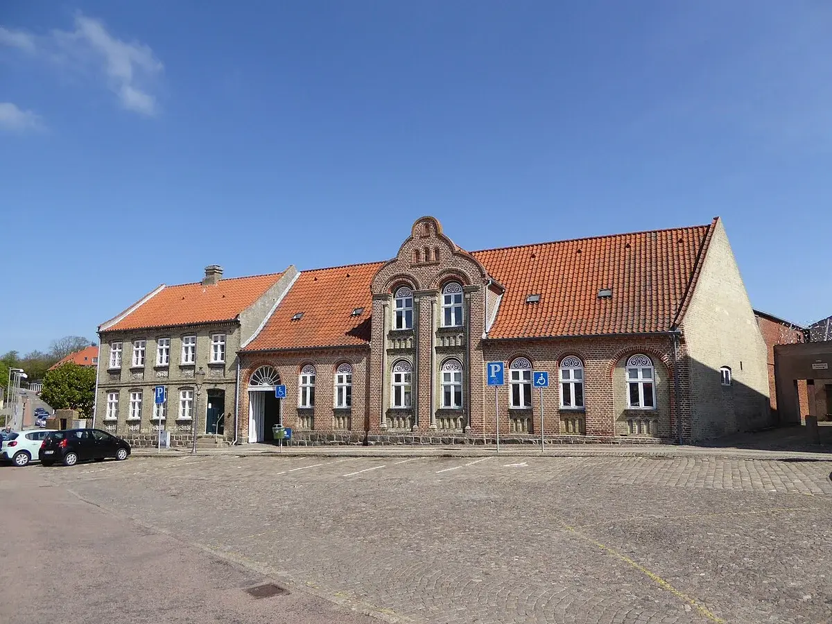 A historic-looking building on a paved square with parking signs and parked cars.