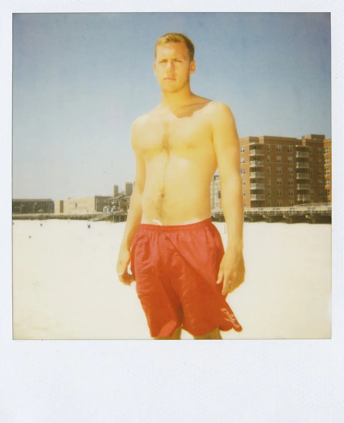 Shirtless lifeguard standing on a sunny beach wearing red shorts