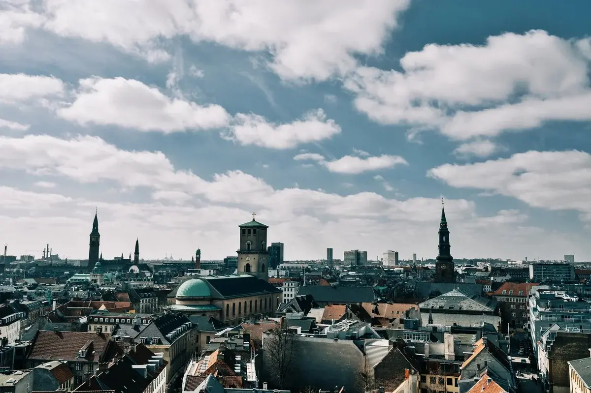 Aerial view of Copenhagen skyline with church spires and rooftops under a cloudy blue sky.
