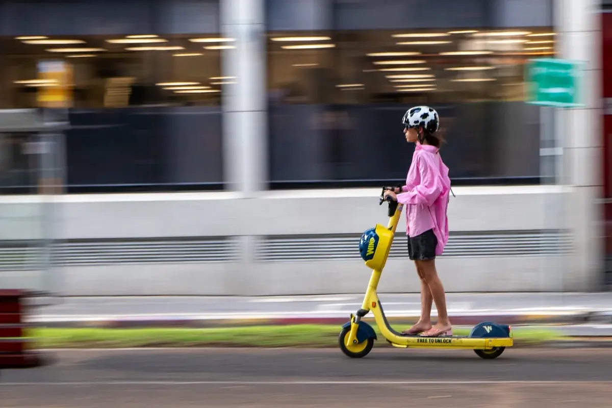 Woman in a pink jacket riding a yellow electric scooter on a city street