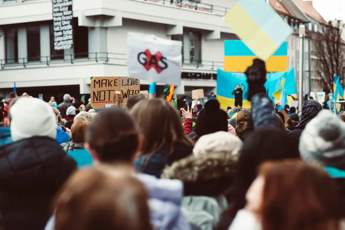A crowd at an anti-war protest holds Ukrainian flags and handmade signs in a city street.