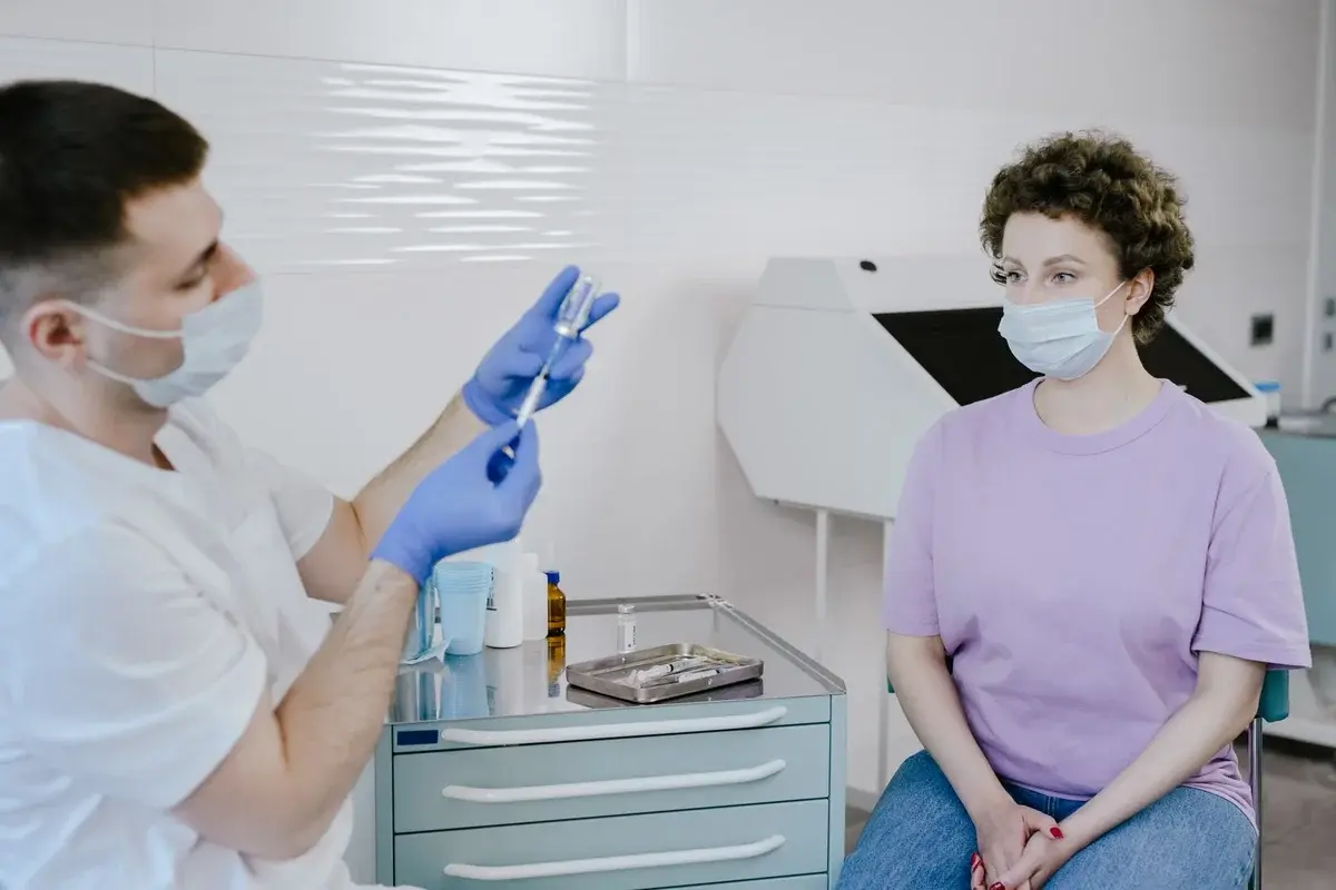 Masked doctor preparing a vaccine shot for a seated patient in a clinic.