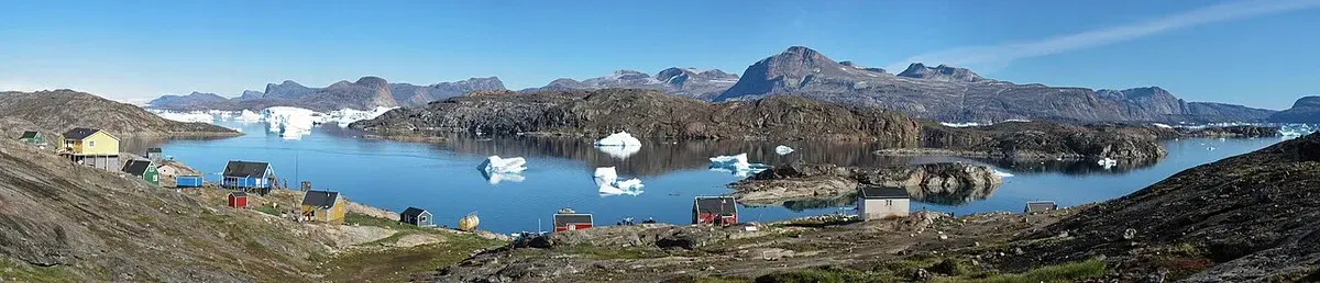 Panorama of a small Greenlandic settlement with houses, water, icebergs, and mountains.