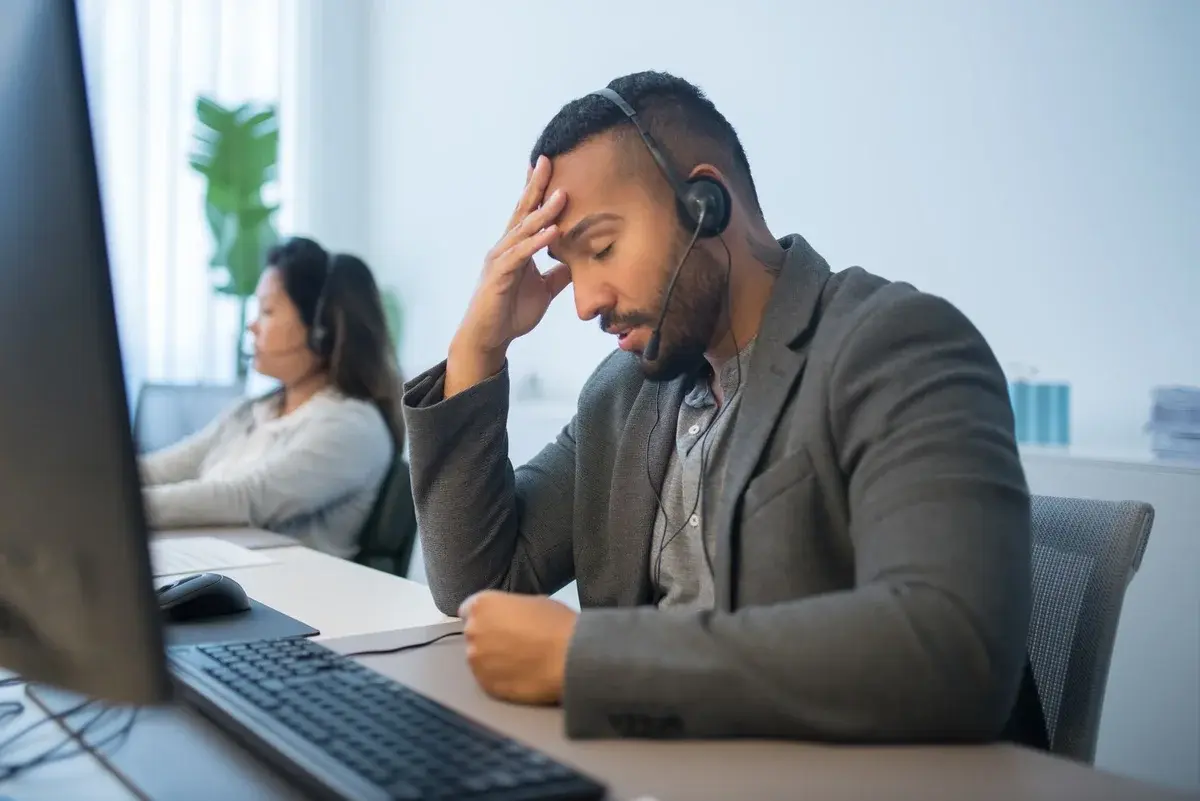 A stressed call center worker with a headset sits at his desk in an office.