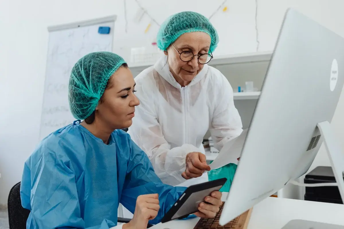 Two female researchers in protective clothing looking at data on a tablet and paper in a lab