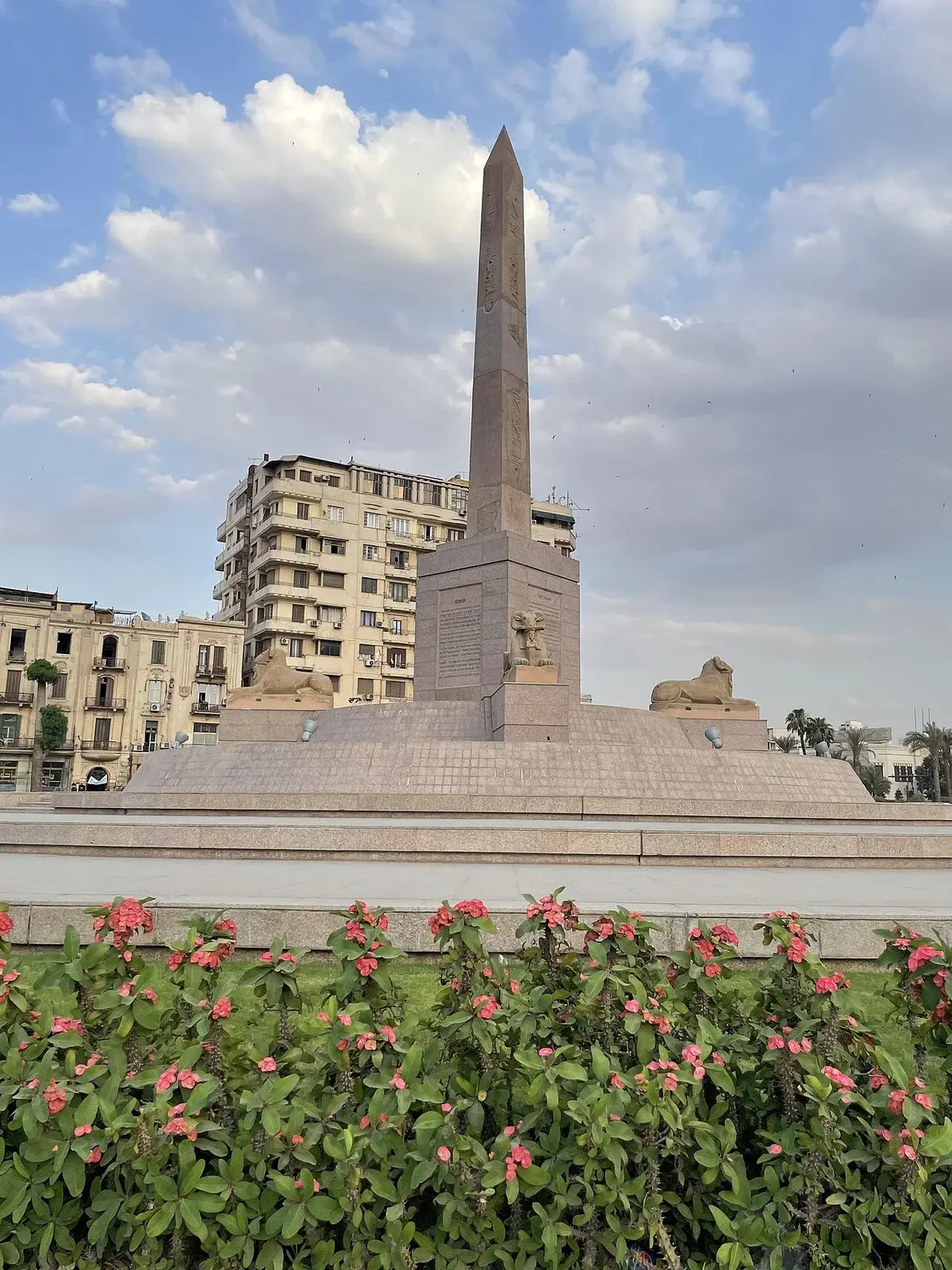 An obelisk monument in Tahrir Square, Cairo, with sphinx statues and city buildings behind it.