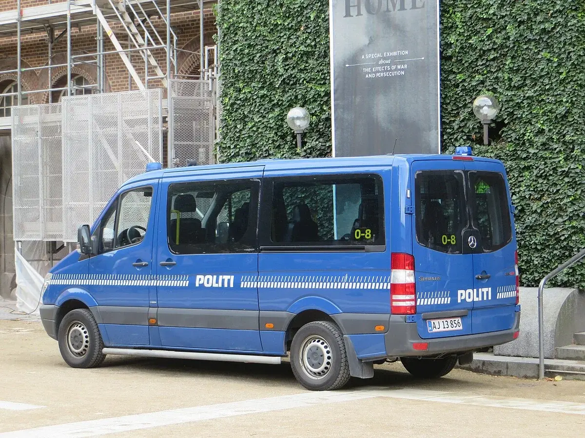 A blue Danish police van marked POLITI parked beside an ivy-covered building with scaffolding.