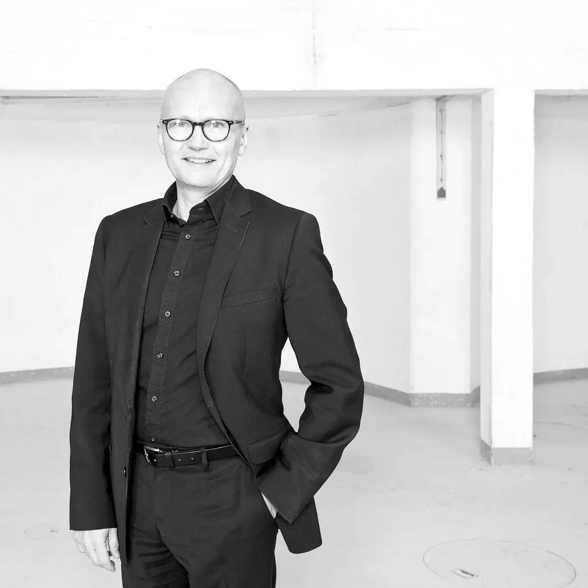 Black-and-white portrait of a man in a suit and glasses standing indoors.