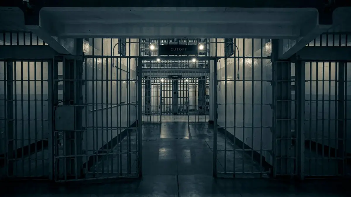 A dim prison corridor with barred gates and cell blocks.
