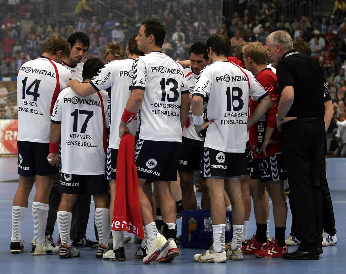 Handball team huddle during a timeout in an indoor arena.