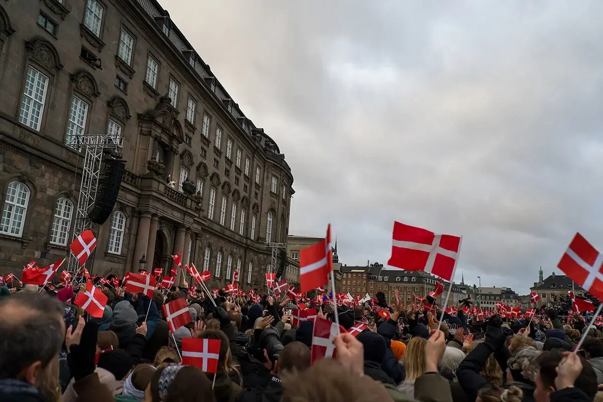 Crowd waving Danish flags outside a palace during a royal proclamation.