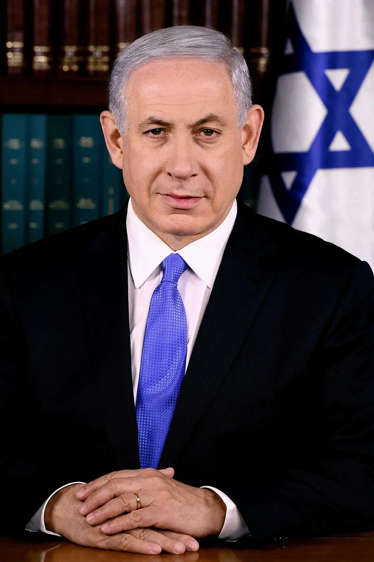 Formal portrait of Benjamin Netanyahu seated in front of bookshelves and an Israeli flag.