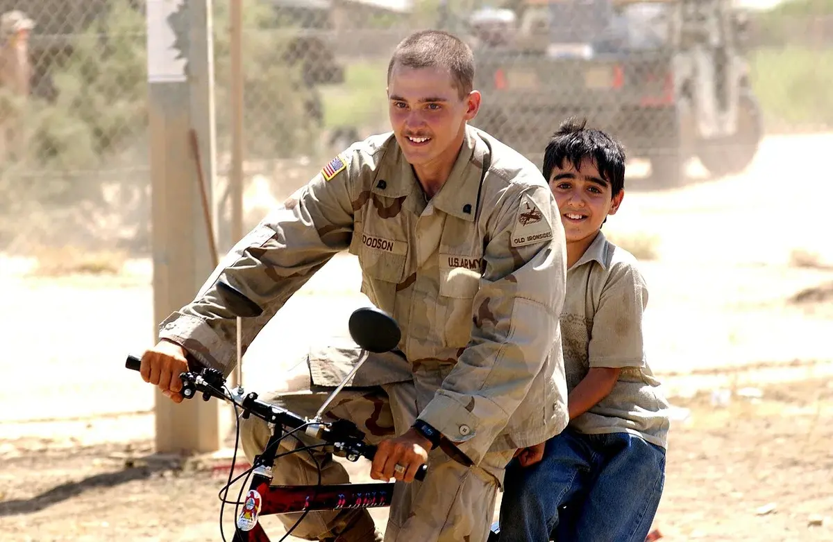 An American soldier rides a bicycle with an Iraqi boy sitting behind him.