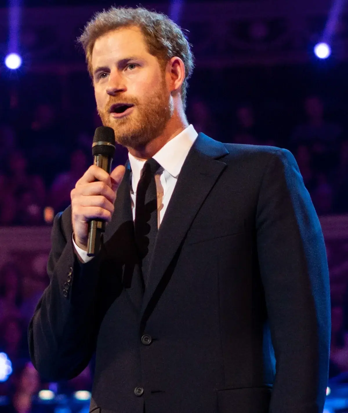 Prince Harry speaking into a microphone on stage in a dark suit.