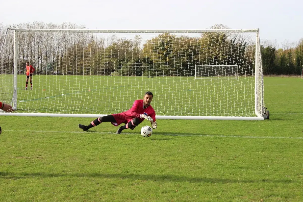 A goalkeeper dives to save a soccer ball near the goal on a grassy field.