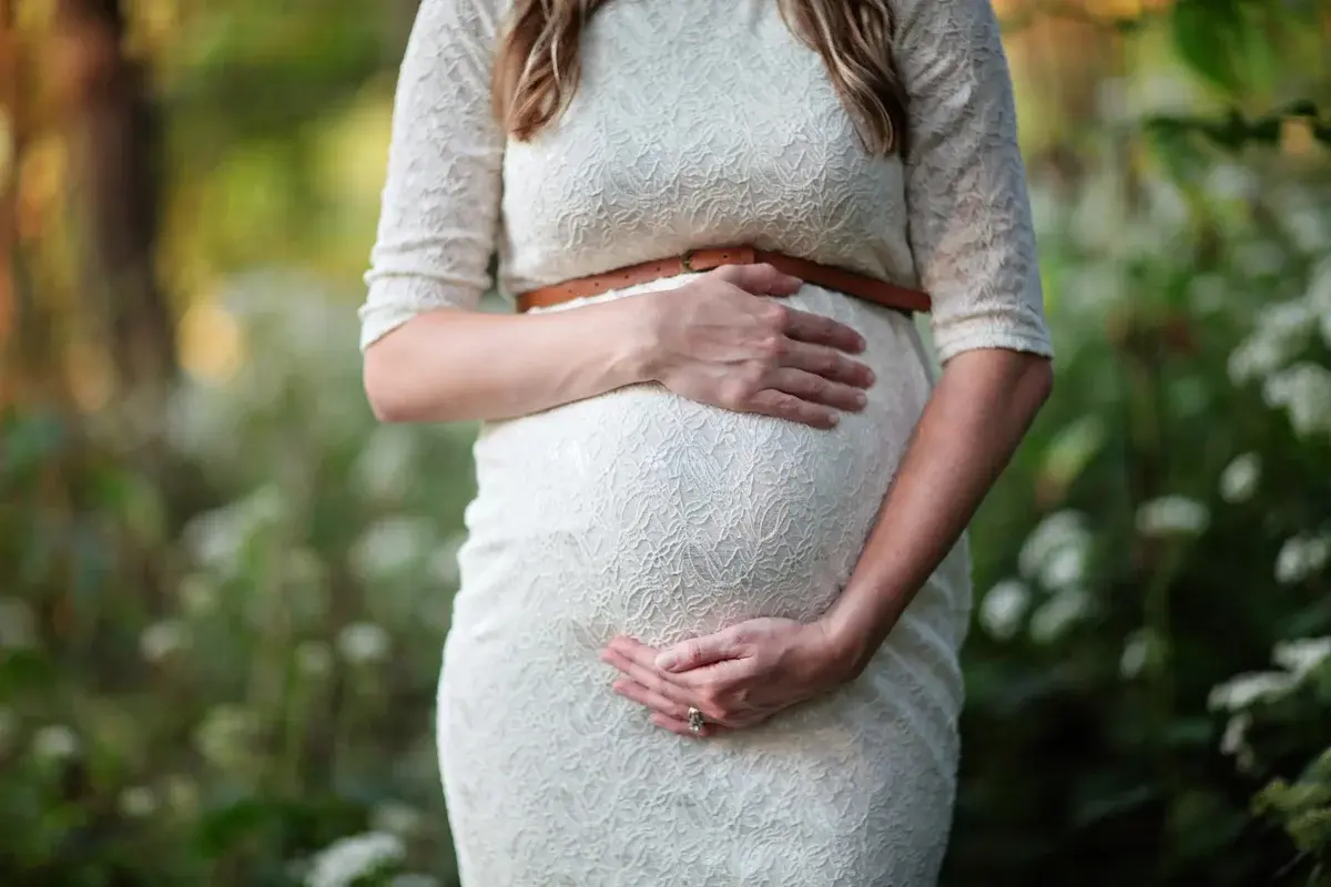 Pregnant woman in a white lace dress holding her belly outdoors.