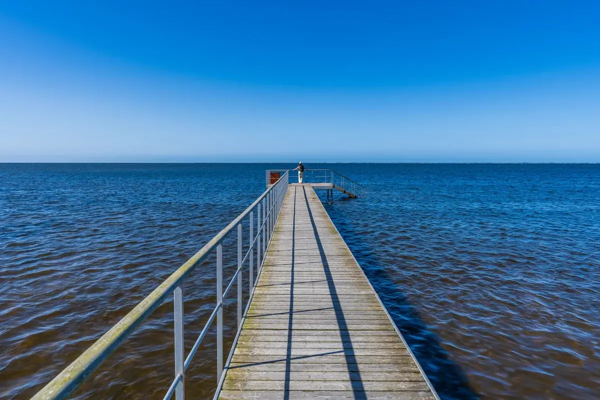 A wooden pier extends into a blue sea with one person standing at the far end.