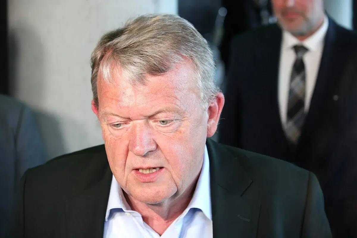 Older man in suit speaking indoors, with another man in the background