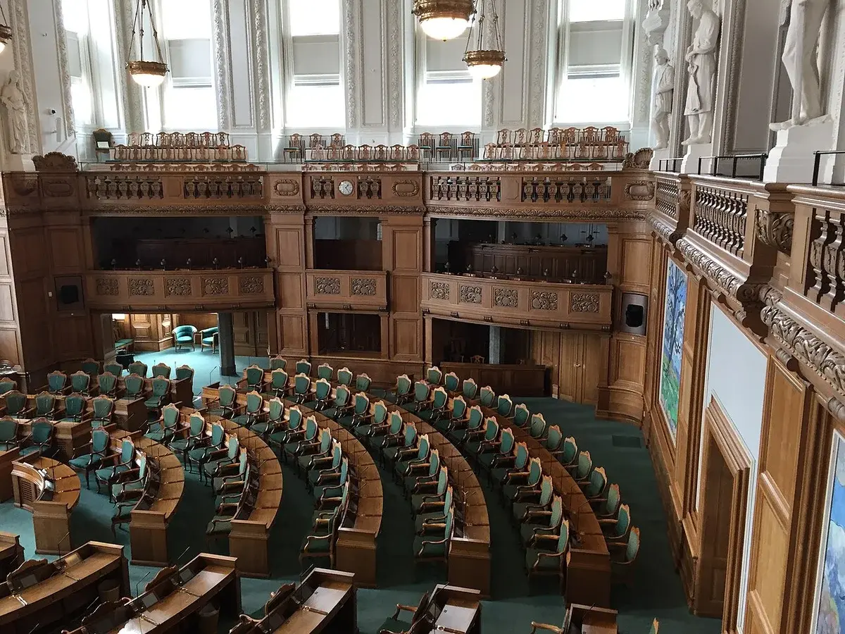 Interior of the Danish parliament chamber with curved rows of desks and green chairs.