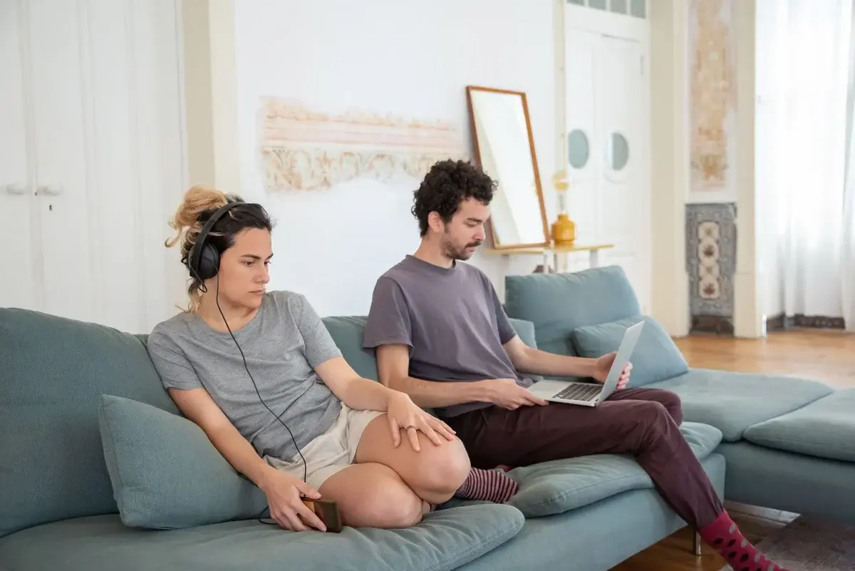 A couple sitting on a sofa at home, one wearing headphones and the other using a laptop.