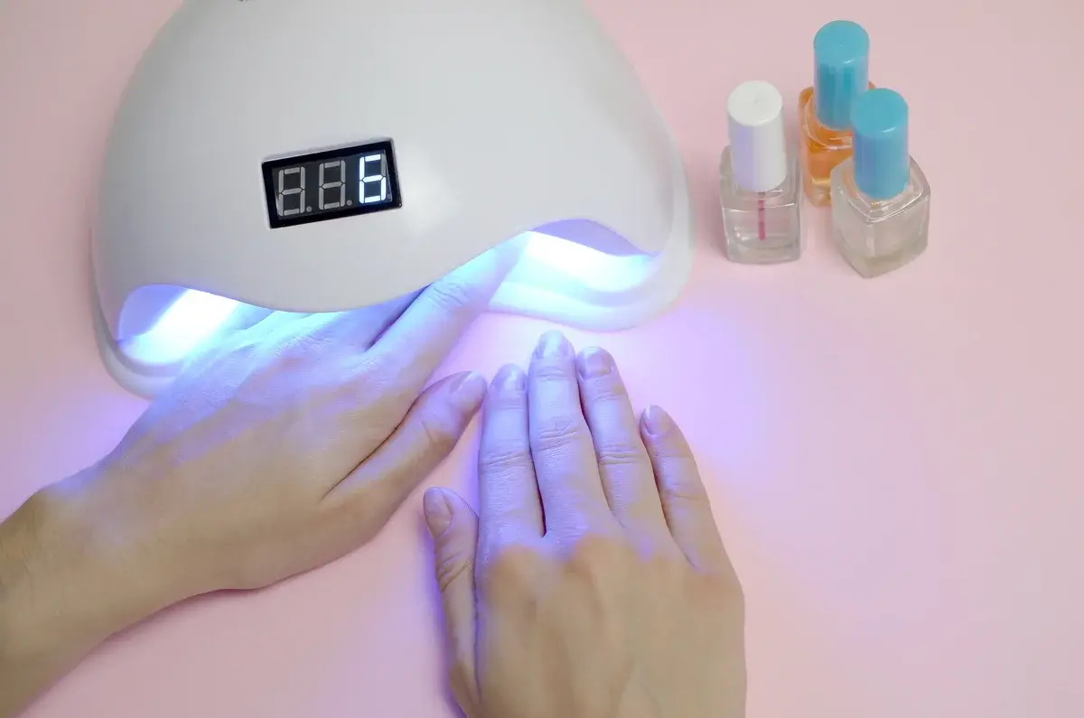 Hands under a manicure lamp on a pink background with nail polish bottles.