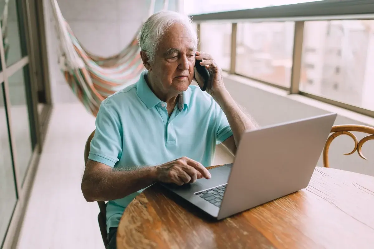 An older man uses a laptop while talking on a smartphone at home.