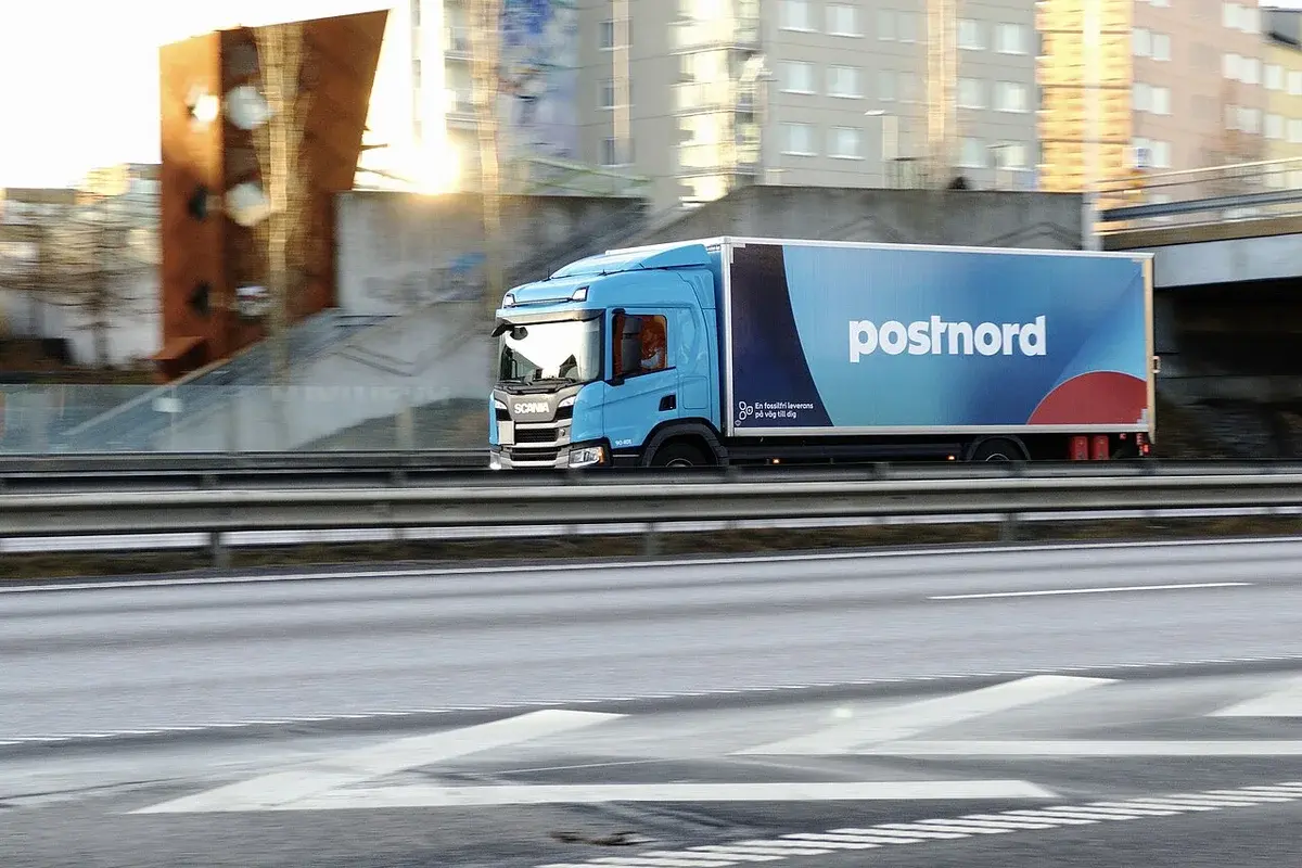 A blue PostNord delivery truck drives past buildings on an urban road.