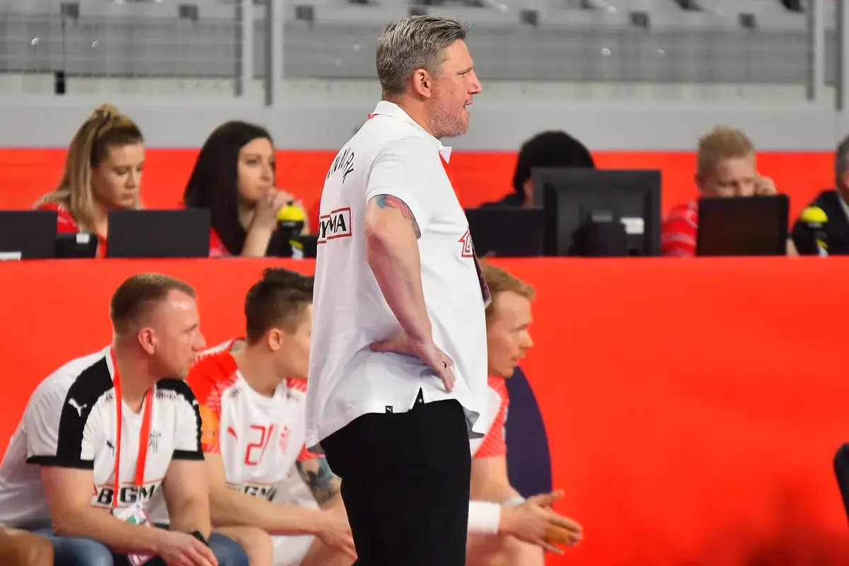 A handball coach stands on the sideline during a match with players seated behind him.