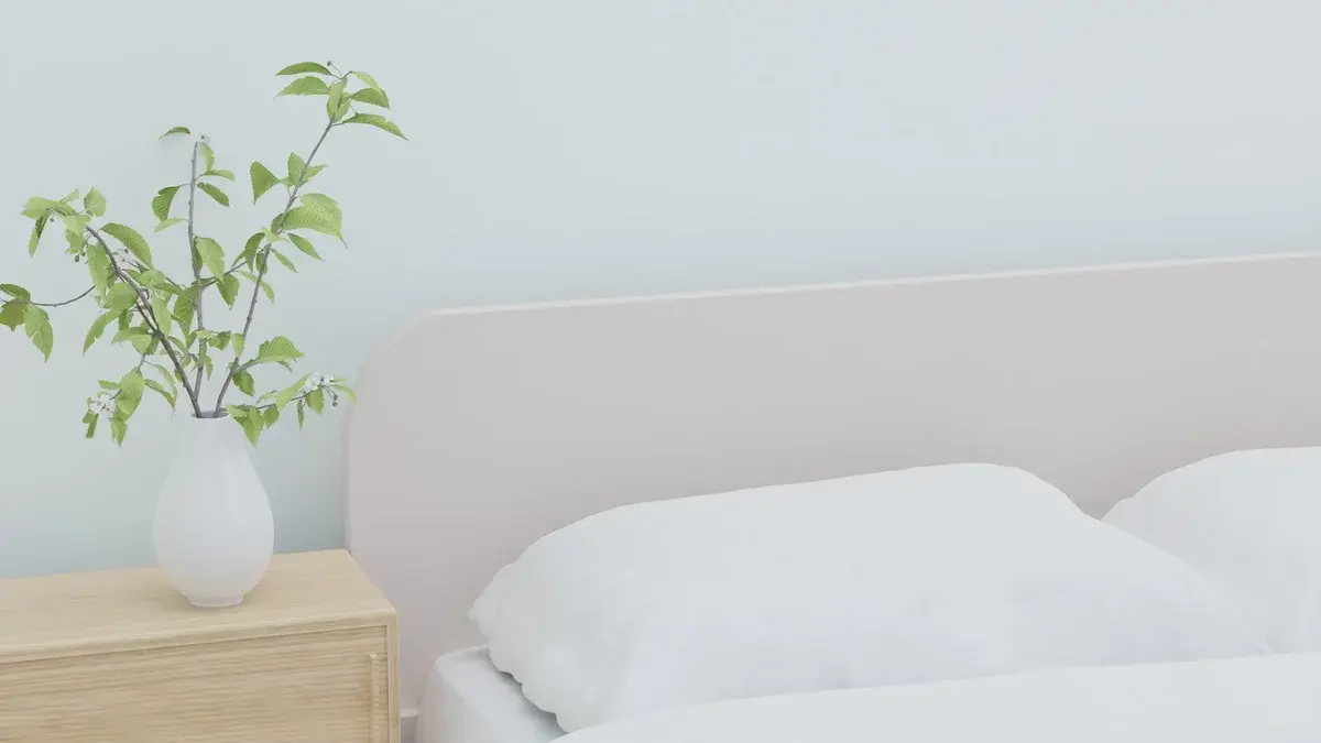 A bright bedroom with white bedding and a vase of green branches on a wooden nightstand.