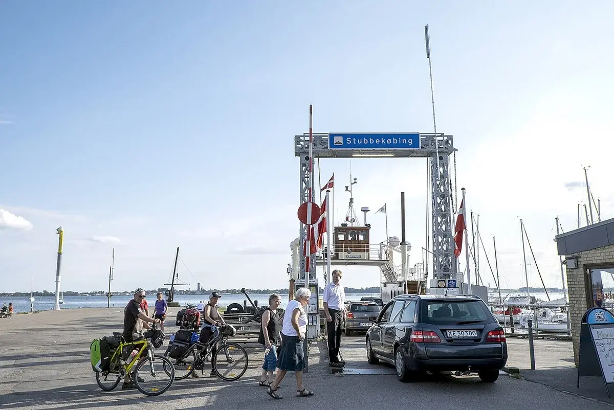 A small ferry at the Stubbekøbing terminal with cars, cyclists, and pedestrians nearby.