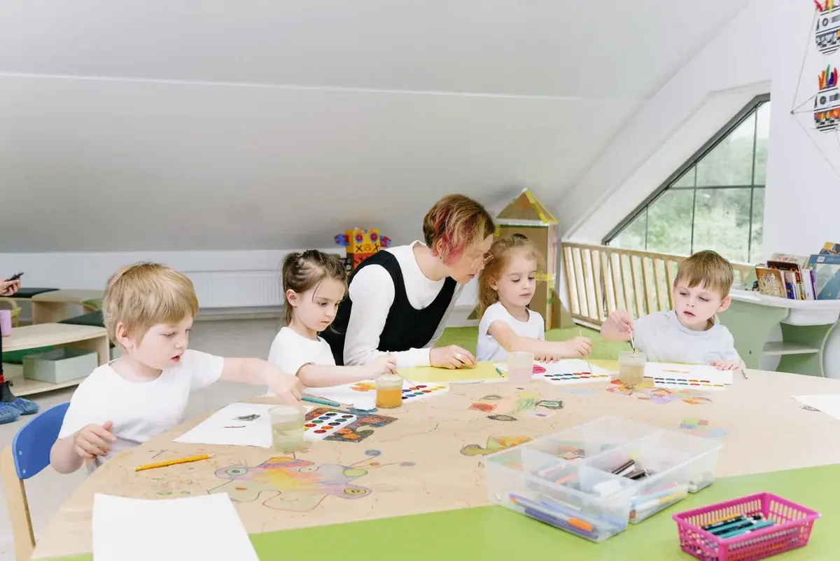 Teacher and young children doing art activities at a table in a bright kindergarten classroom.