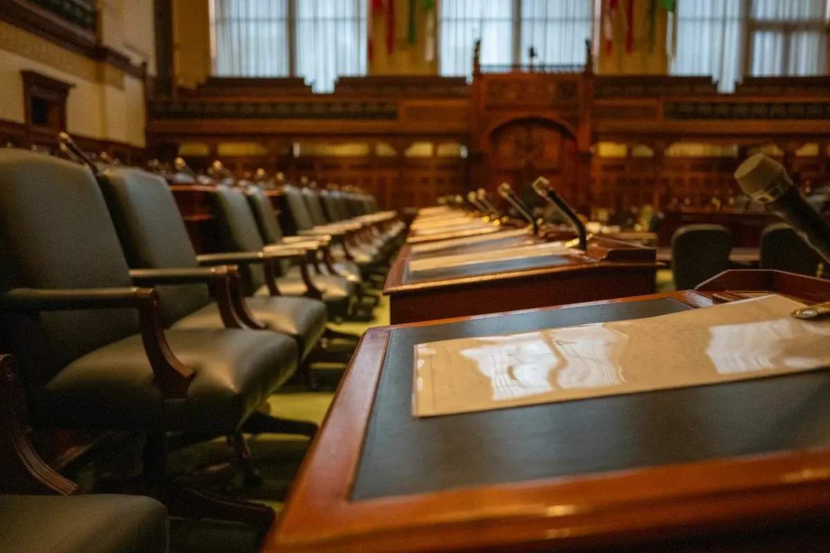 Empty legislative chamber with desks and microphones in a wood-paneled room.