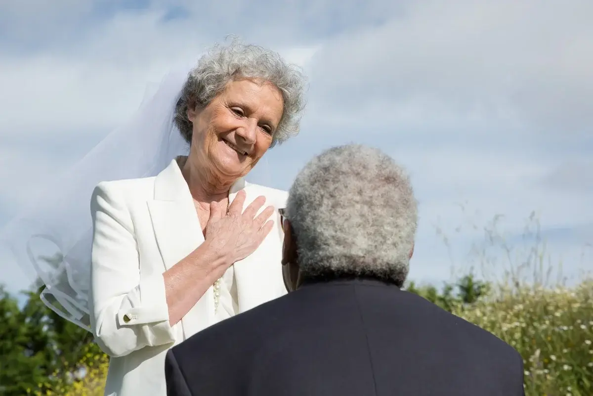 An elderly bride smiles at her groom during an outdoor wedding ceremony.