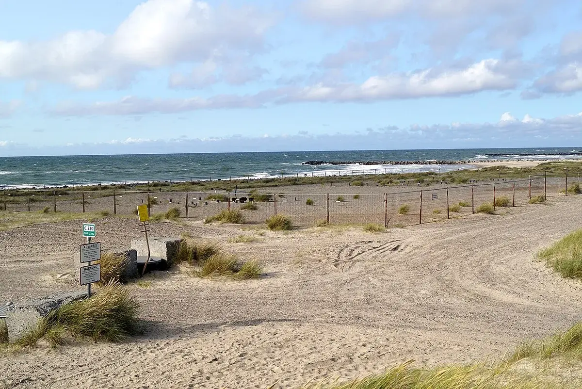 Fenced chemical depot area on a sandy coastal landscape with warning signs and the sea beyond.