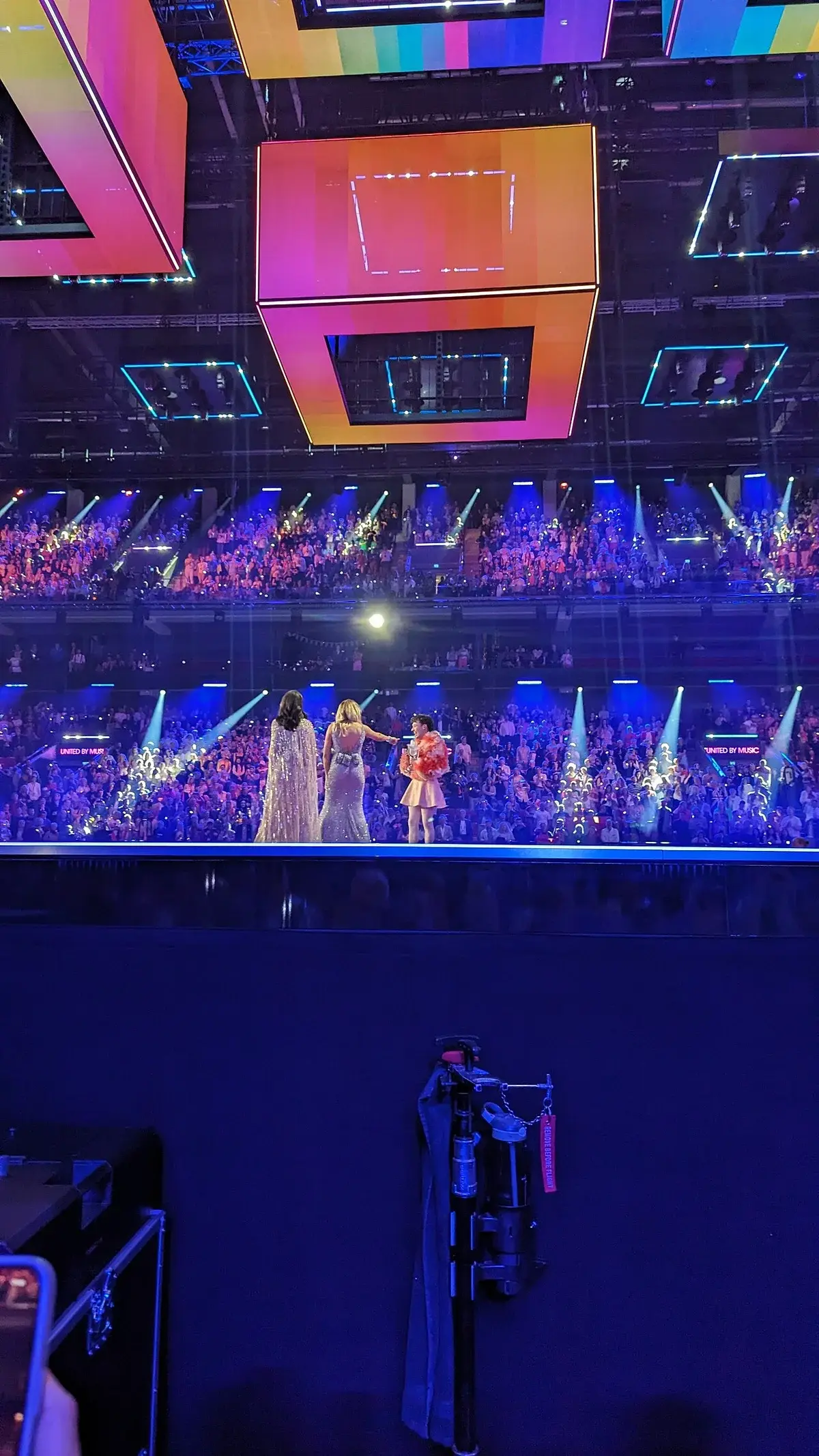 Nemo stands on stage with two hosts in a packed Eurovision arena.